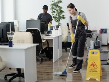 Young Black Man Wiping Computer Monitors While Woman With Mop Cleaning Floor
